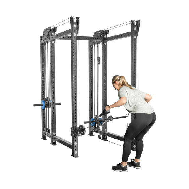 A woman in a gray shirt and black leggings is focused on using a Bells of Steel "Kraken Rack Attachment for 6 Post Manticore Racks" to perform a bent-over row. The sturdy black equipment supports her strength training routine with dual-handle cables.