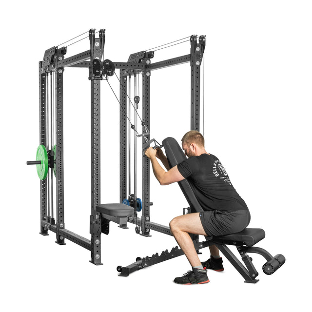 A man works out on a Bells of Steel Kraken Rack Attachment for 6 Post Hydra Racks, pulling cables from the compact system with weight plates while seated on an incline bench. He's wearing a black shirt and shorts against a plain white background.