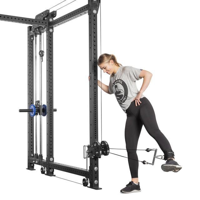 A woman in a gray t-shirt and black leggings performs a leg exercise using the Bells of Steel Kraken Rack Attachment for 6 Post Manticore Racks. She holds onto the frame for support while extending one leg back, enjoying efficient and versatile workouts with adjustable weights and pulleys.