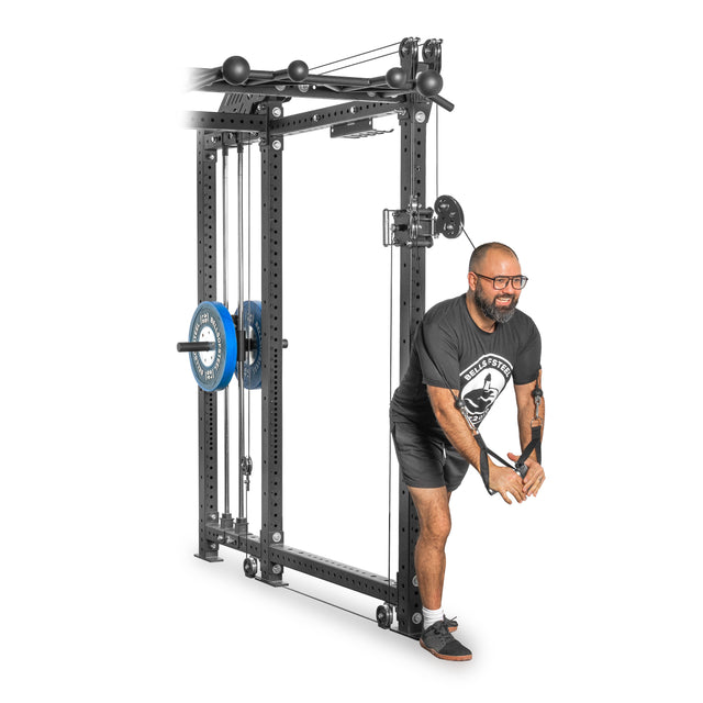 A person works out in a gym using the Kraken Rack Attachment for 6 Post Hydra Racks by Bells of Steel. They're extending their arms downwards while holding cable handles, dressed comfortably in a t-shirt, shorts, and athletic shoes.