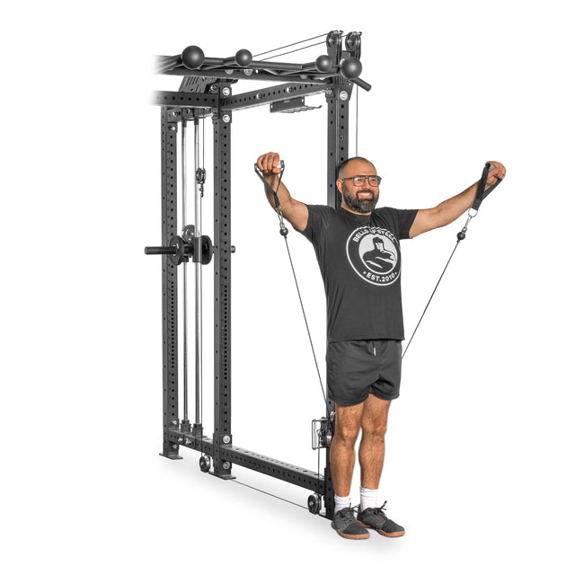 A man, dressed in a black t-shirt, shorts, and glasses, performs a chest fly with Bells of Steel's Kraken Rack Attachment on a Hydra Rack. He stands with feet shoulder-width apart, pulling the cables outward from the sleek attachment resembling a dual-handle cable tower.