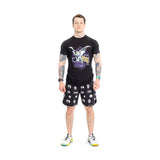 A tattooed man stands against a white backdrop, wearing a black T-shirt with a colorful design, Bells of Steel gym shorts featuring white patterns on stretchy fabric, and vibrant sneakers. His arms are relaxed at his sides as he looks straight ahead.