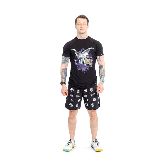 A tattooed man stands against a white backdrop, wearing a black T-shirt with a colorful design, Bells of Steel gym shorts featuring white patterns on stretchy fabric, and vibrant sneakers. His arms are relaxed at his sides as he looks straight ahead.