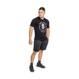 A man in a black athletic T-shirt with white text and Bells of Steel high-performance polyester gym shorts pairs the look with black sneakers and dark socks against a plain white background.