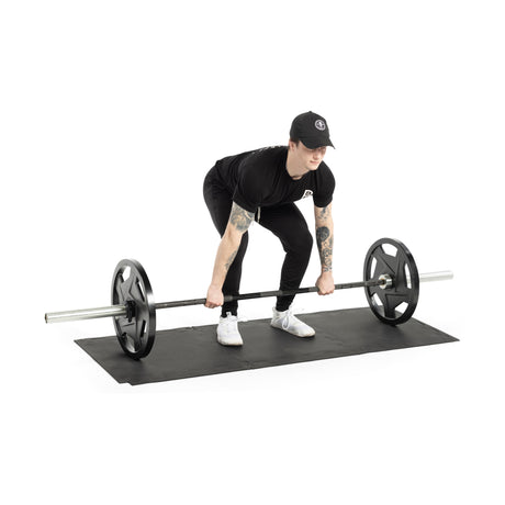 Dressed in black and wearing a cap, a person prepares to lift a barbell on Bells of Steel's 24" x 24" Puzzle Mats, capturing the commercial gym vibe. In a squat position, they grip the barbell with both hands for optimal floor protection.