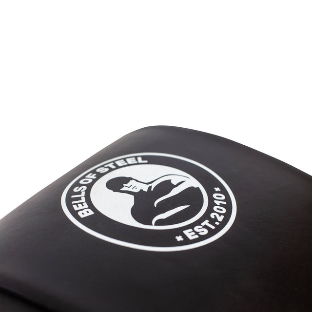 A close-up of a black surface features a white logo with a stylized person lifting weights, surrounded by "Bells of Steel" and "Est. 2010." The emblem is centered on the Sit Up Mat, evoking its design against a plain white background.