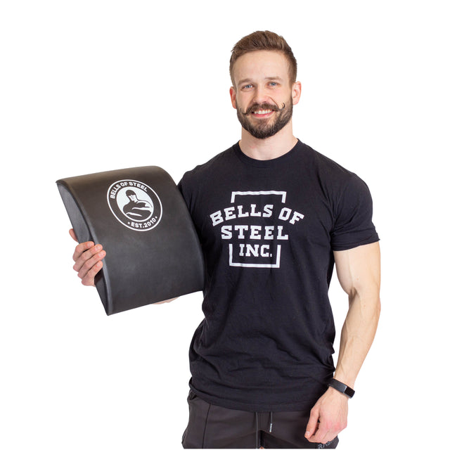 A bearded man in a "Bells of Steel" T-shirt smiles while holding a Sit Up Mat featuring the company's logo. Celebrated for its portability and lumbar support, he stands against a plain white background.