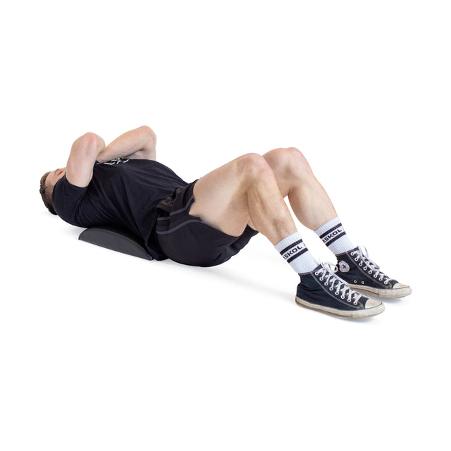 On a Bells of Steel Sit Up Mat, a person lies with knees bent for lumbar support, feet flat. They're wearing a black shirt and shorts, paired with chic black high-tops and white socks. The spotless white background accentuates their fitness routine.