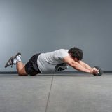A man in a gray t-shirt and black shorts uses the Bells of Steel USA ABsyidyan Stainless Steel Ab Roller, kneeling on a gym floor as he performs an ab rollout with outstretched arms against a plain background.