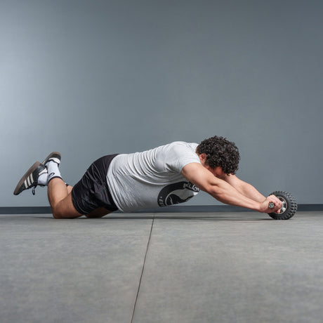 A man in a gray t-shirt and black shorts uses the Bells of Steel USA ABsyidyan Stainless Steel Ab Roller, kneeling on a gym floor as he performs an ab rollout with outstretched arms against a plain background.