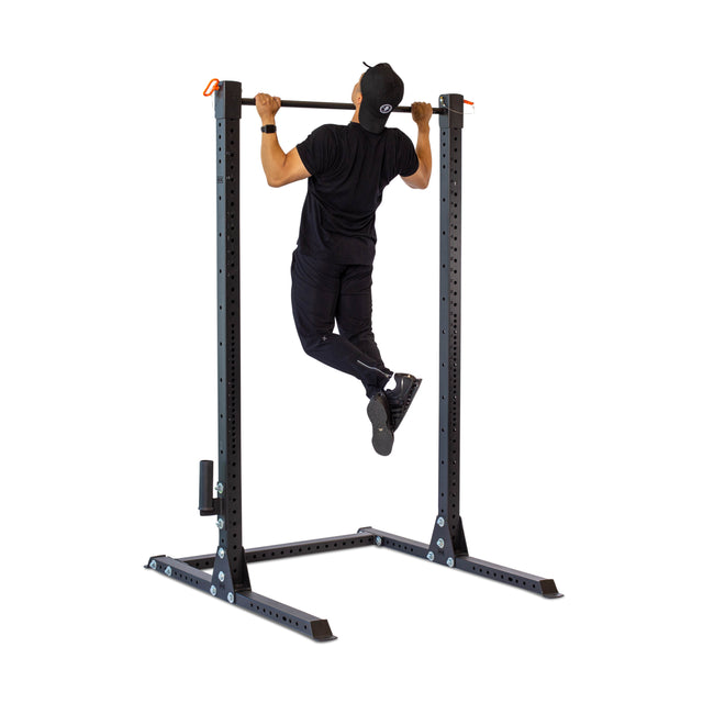 A man in a black outfit and cap uses the Bells of Steel Adjustable Pull-up Bar Rack Attachment with a sturdy base for customizable height settings, demonstrating its versatility as essential workout equipment in a well-lit, neutral space.