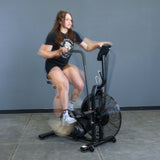 A woman in a black shirt and shorts pedals quickly on the Bells of Steel Residential Air Bike with Wind Guard, creating motion blur as she works out intensely indoors against a gray wall.