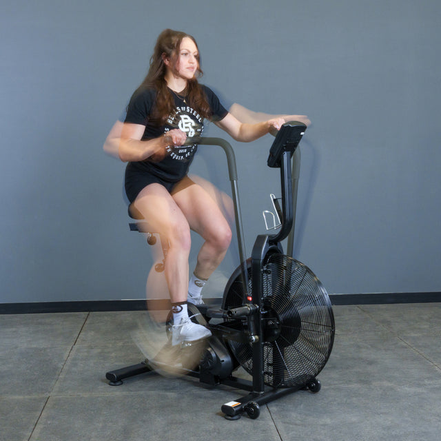 A woman in a black shirt and shorts pedals quickly on the Bells of Steel Residential Air Bike with Wind Guard, creating motion blur as she works out intensely indoors against a gray wall.