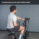 A user sits on the Bells of Steel Residential Air Bike with Wind Guard, reaching for the phone holder on the handlebars. Text above says, "Includes phone and water bottle holder." A plain gray wall sets a home gym vibe.