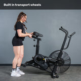 A woman in athletic wear tilts the Bells of Steel Residential Air Bike with Wind Guard onto its front wheels for easy moving. Above her, the caption reads, "Built in transport wheels." Shown indoors, it’s the ideal addition to any home gym.