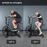 Two people, 6'2" and 5'1", ride Bells of Steel Residential Air Bikes with Wind Guard side by side at home. Text reads "Suitable for all heights." Both look comfortable and well-positioned on the bikes.