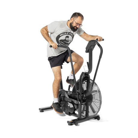 A man in a gray T-shirt and black shorts exercises on a Bells of Steel Residential Air Bike with Wind Guard, perfect for a full-body workout. He has short hair and glasses. The bike includes a digital display and handlebars, all set against a plain white background in his home gym.