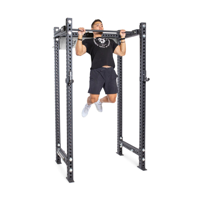 A person uses a Bells of Steel Adjustable Pull-up Bar Rack Attachment on a sturdy power rack, wearing black T-shirt, shorts, and white sneakers. The bar features multiple adjustment holes and a robust frame.