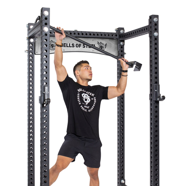 A man demonstrates how to attach an Adjustable Pull-up Bar Rack Attachment by Bells of Steel to a metallic power rack, featuring multiple holes for custom adjustment. The black-clad man focuses on securing the bar for optimal stability.