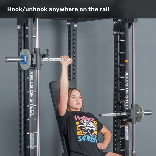 A woman in a graphic tee uses Anvil Handles by Bells of Steel USA on a Smith Machine, pressing a barbell overhead while seated. Text above reads, “Hook/unhook anywhere on the rail.”.