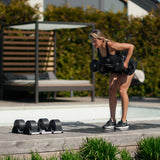 A woman in athletic wear performs a bent-over row with Nuobell NÜOBELL Adjustable Dumbbells on a wooden deck outdoors, surrounded by machined steel weight plates, with a shaded lounge area in the background.