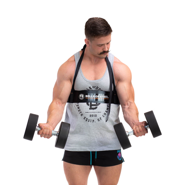 In a plain white studio, a muscular man with a mustache performs bicep curls using dumbbells, maintaining proper form. He wears a gray tank top and black shorts, and for added support, an aluminum Arm Blaster by Bells of Steel.