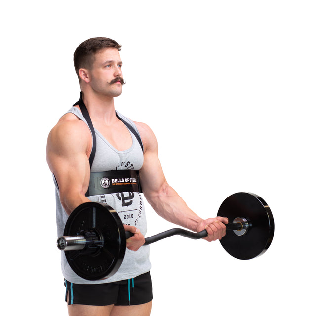 A mustached man in a tank top and shorts performs a barbell curl with a Bells of Steel Arm Blaster for proper form, using lifting straps for support, against a plain white background.