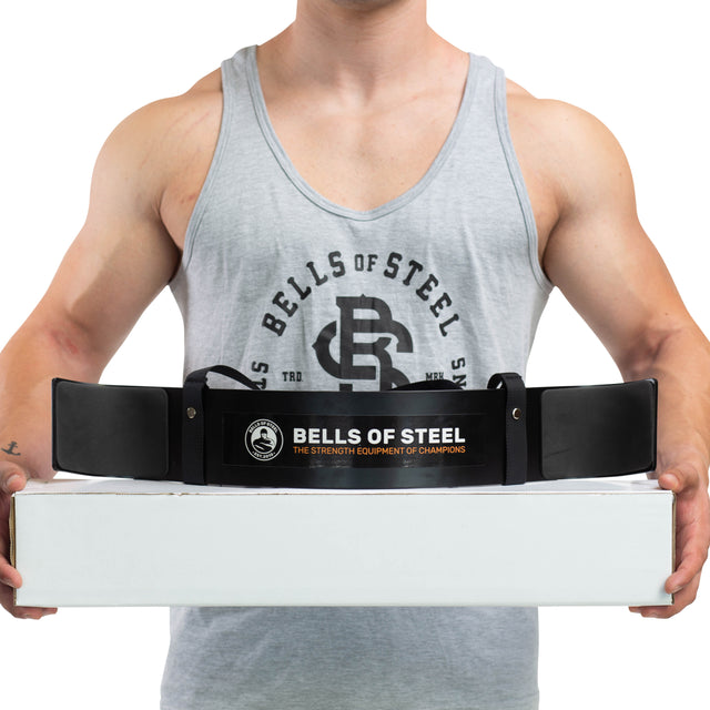 A person in a gray tank top holds a white box with "Bells of Steel" on a black belt draped over it, featuring an Arm Blaster made from durable aluminum for optimal form.