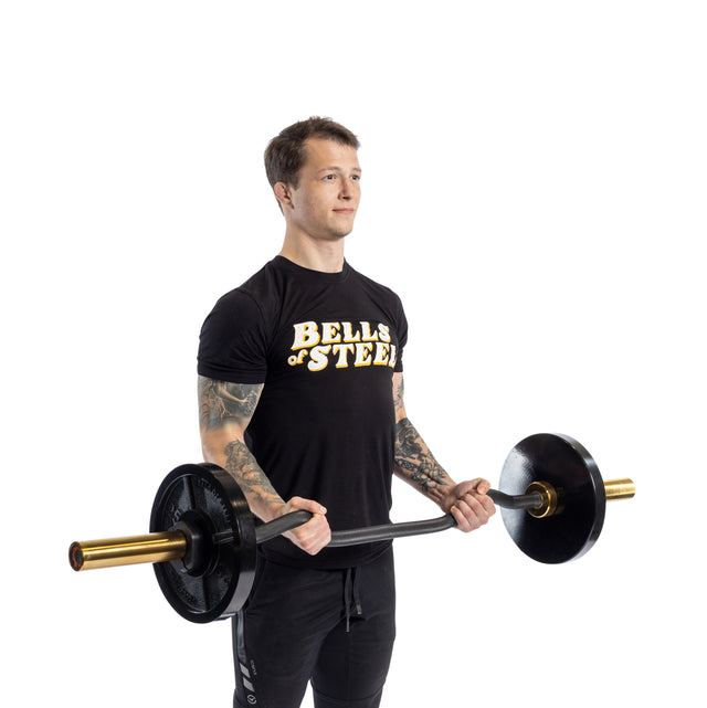 A tattooed person in a "Bells of Steel" t-shirt demonstrates impressive arm development while performing bicep curls with the Bells of Steel EZ Curl Bar (54.5") against a plain white background, gripping it with both hands and bending at the elbows.