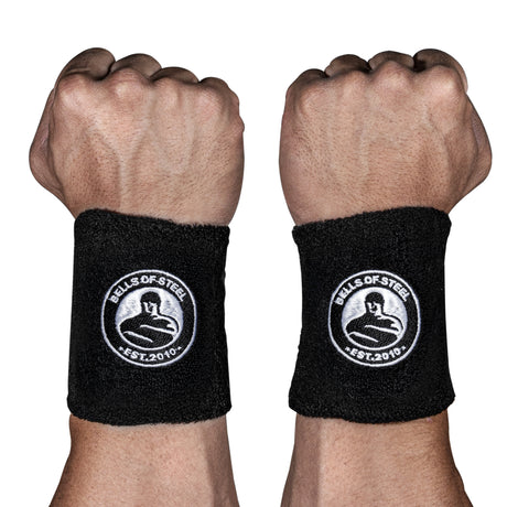 Two fists with wrists facing forward, each wearing Bells of Steel BellGuard Kettlebell Wrist Guards—black guards bearing a circular kettlebell logo and "EST. 2010" in white—essential workout accessories.