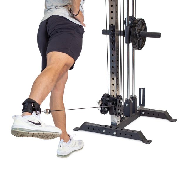 A person performs a leg kickback exercise with an Adjustable Ankle Strap Cable Attachment from Bells of Steel, emphasizing glute training. They're in athletic shoes, shorts, and a gray shirt, showcasing the interaction between their leg and the machine.