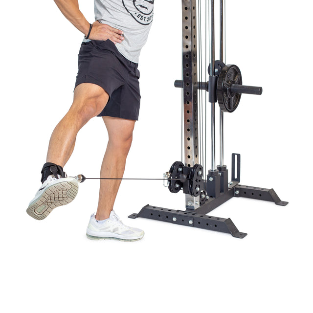 Wearing a gray shirt, black shorts, and white sneakers, a person skillfully uses the Bells of Steel Adjustable Ankle Strap Cable Attachment to focus on glute training. They extend one leg to pull the cable on the weight-equipped machine effectively.