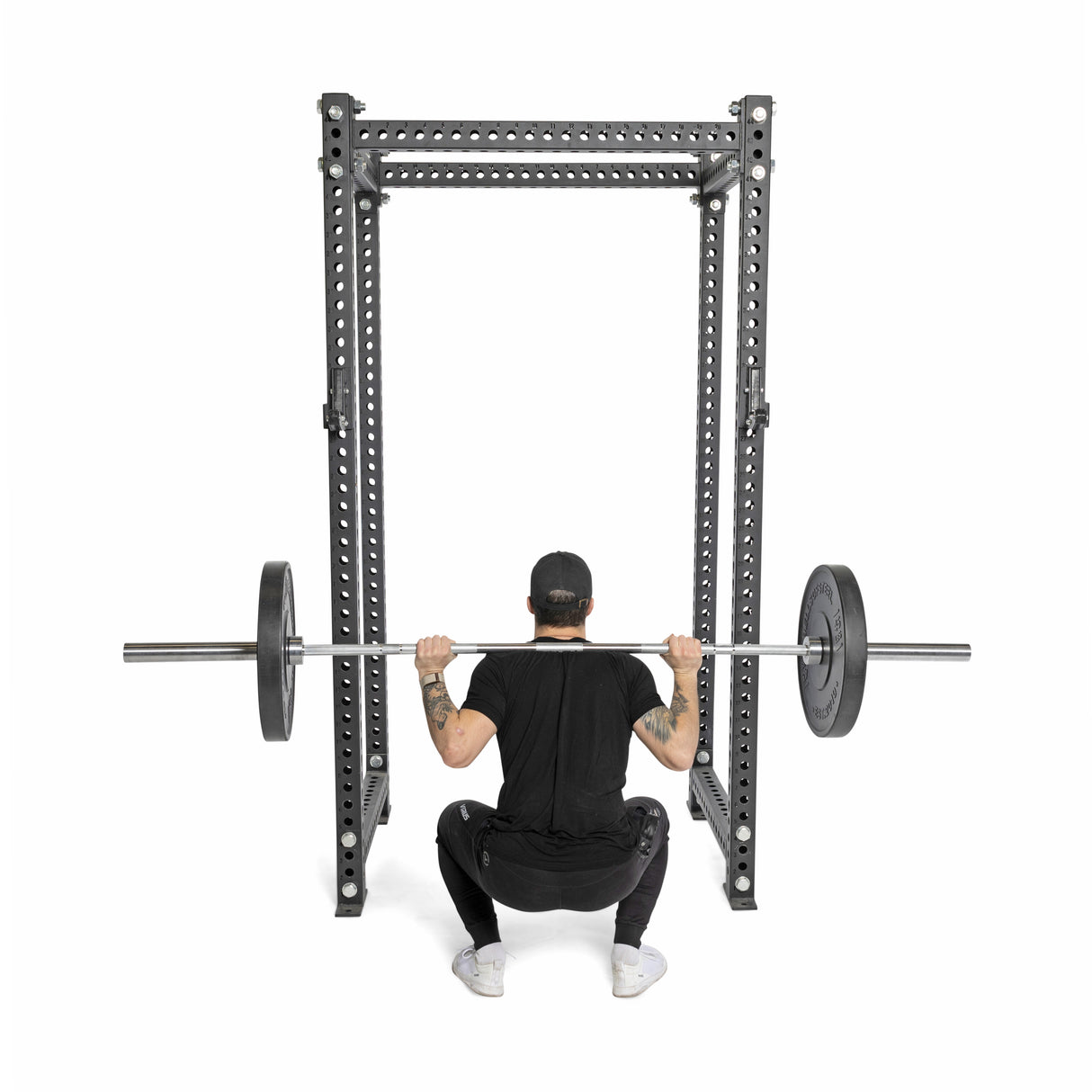 Wearing athletic gear, a person performs a barbell squat with precision using Bells of Steel's All-Black Bumper Plates. They face away on a squat rack, set against a white backdrop that emphasizes the focus and intensity of their workout.