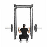 Wearing black workout gear and a cap, an individual performs a barbell squat with Bells of Steel All-Black Bumper Plate Sets inside a metal power rack, viewed from behind on a white background for safe lifting.