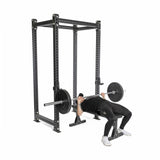 Dressed in black workout clothes and white sneakers, a person demonstrates safe lifting inside a power rack, bench pressing a barbell fitted with Bells of Steel All-Black Bumper Plate Sets against a plain white background.