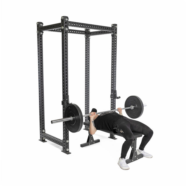 Dressed in black workout clothes and white sneakers, a person demonstrates safe lifting inside a power rack, bench pressing a barbell fitted with Bells of Steel All-Black Bumper Plate Sets against a plain white background.