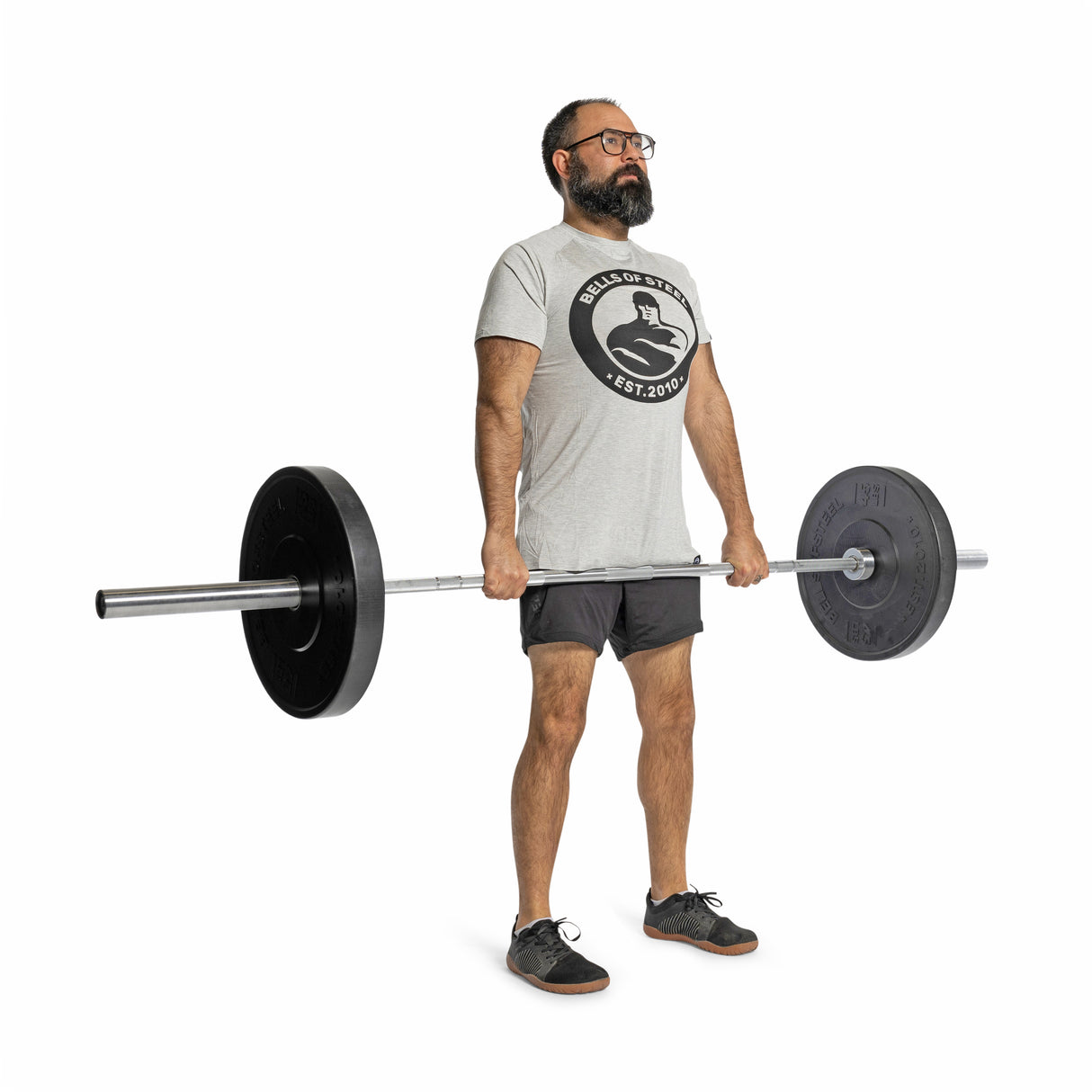 A bearded man in glasses, a gray t-shirt, and shorts lifts a barbell with Bells of Steel All-Black Bumper Plates against a white background, gripping the bar firmly.