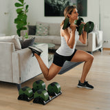 A woman in athletic wear does a Bulgarian split squat in her living room using Nuobell NÜOBELL Adjustable Dumbbells, with one foot on the sofa and several machined steel weight plates arranged neatly nearby.