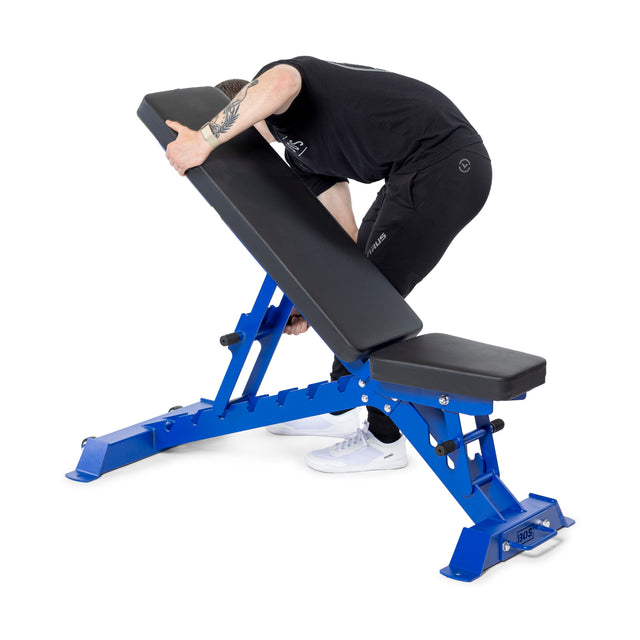 Wearing a black outfit and white sneakers, a person adjusts the Buzz-Saw Heavy-Duty Adjustable Bench from Bells of Steel, tilting its blue and black backrest forward against the white background.