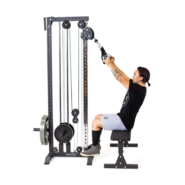 A person in a black t-shirt, gray shorts, and black shoes uses the Bells of Steel Cable Tower for a workout. Seated on a bench with short hair, they skillfully engage the weight stack while adjustable handles adapt to their routine against a white background.