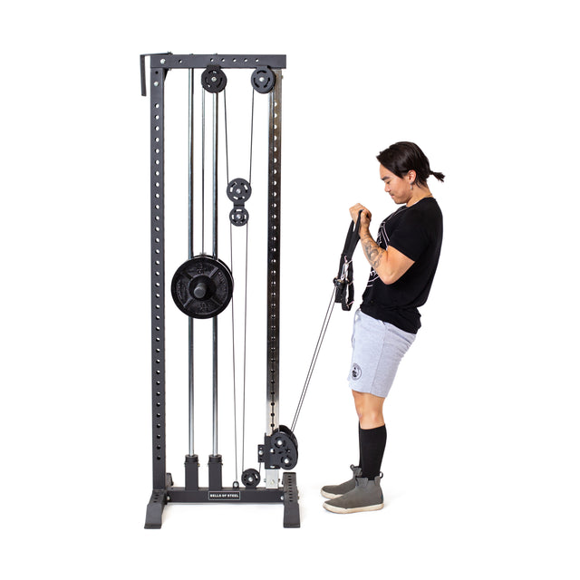 Someone in a black shirt, gray shorts, and sneakers is using a Bells of Steel Cable Tower. They face the tower, pulling handles upwards to their chest for exercise with resistance from the machine's weight stack and pulleys.