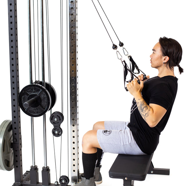 A person in a black t-shirt and gray shorts is using the Bells of Steel Cable Tower on a bench. They pull adjustable handles toward themselves, with a white background accentuating the machine's weight stack.