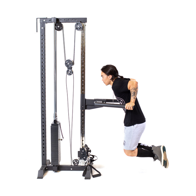 A person in athletic wear performs tricep dips on the Bells of Steel Cable Tower with adjustable handles. Against a plain white background, they wear a black t-shirt, gray shorts, black knee-high socks, and black shoes.