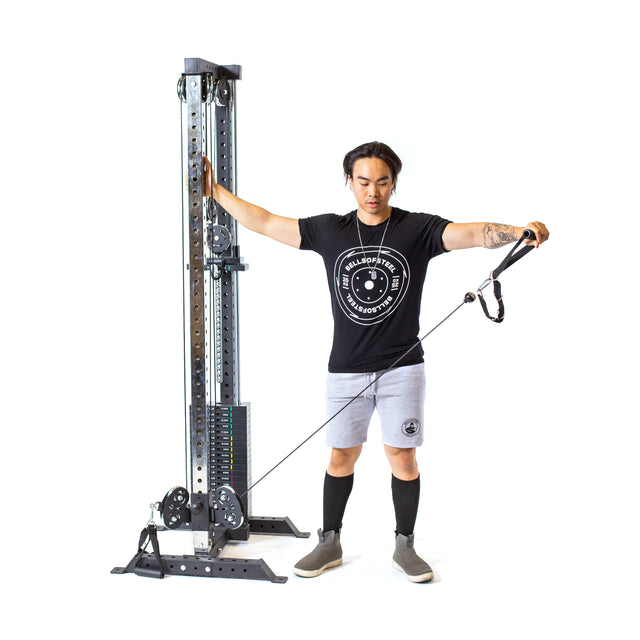 A person at the Bells of Steel Cable Tower grips an adjustable handle with one hand and pulls a cable with the other, wearing a black t-shirt, gray shorts, and long black socks against a plain white background.