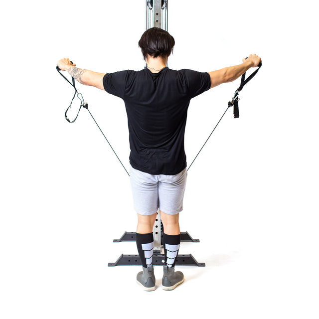 A person in a black shirt, gray shorts, and striped socks does cable exercises on the Bells of Steel Cable Tower. They stand with arms extended sideways, gripping adjustable handles connected to the machine's weight stack against a white background.