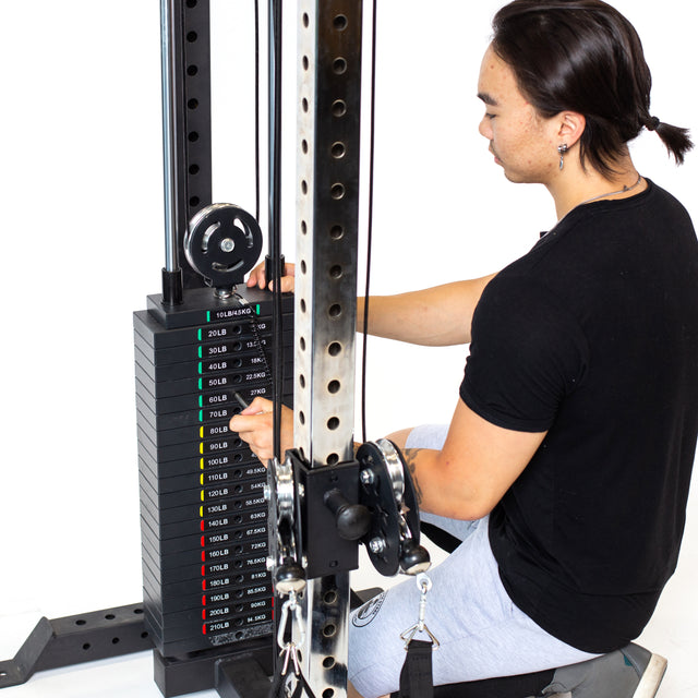 A person in a black shirt adjusts the weight stack on the Bells of Steel Cable Tower. The weights range from 10 to 20 pounds, and the machine has a metal frame with adjustable handles, cables, and pulleys for versatile workouts.