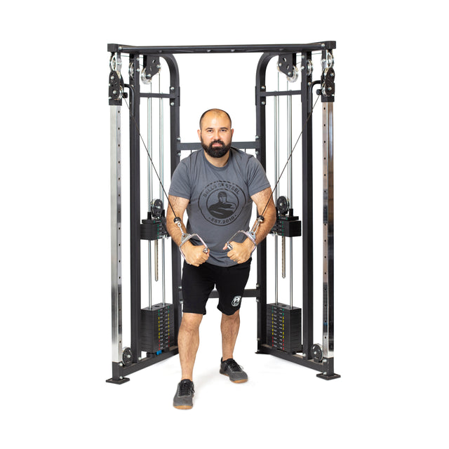 A man intensely focuses as he works out on a Bells of Steel Functional Trainer. In a gray t-shirt and black shorts, he grips the cable attachments with determination. The sleek metal structure has pulleys and adjustable weights designed for versatile workouts.