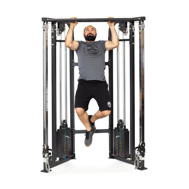 A bearded individual performs a pull-up on a Bells of Steel Functional Trainer, wearing a gray T-shirt, black shorts, and black shoes. The trainer's visible weights and pulleys highlight its versatile cable attachments for an efficient workout.