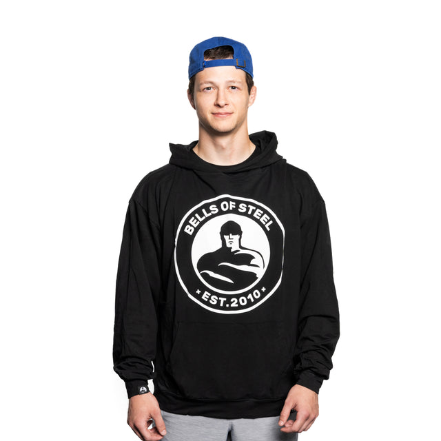 Wearing the Bells of Steel Bamboo Hoodie, featuring a buttery-soft bamboo blend and "EST. 2010" text, a person stands against a white background highlighting their fit figure. They smile slightly while looking at the camera under their blue cap.