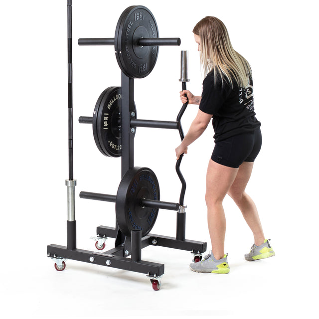 A person in a black shirt and shorts adjusts weights on a Bells of Steel Bumper Plate Weight Tree And Bar Holder. This sleek storage system includes various bars and plates. They stand on a white background, wearing sneakers.